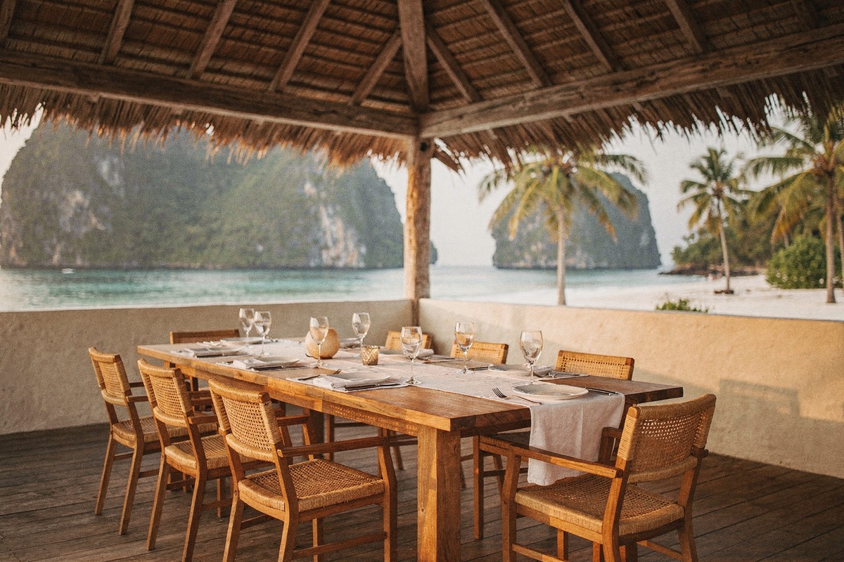 A long timber table set for eight with linen runners, glassware and small vases, cane-backed chairs, under a thatched pavilion on a timber deck, karst headlands, palms and turquoise sea.