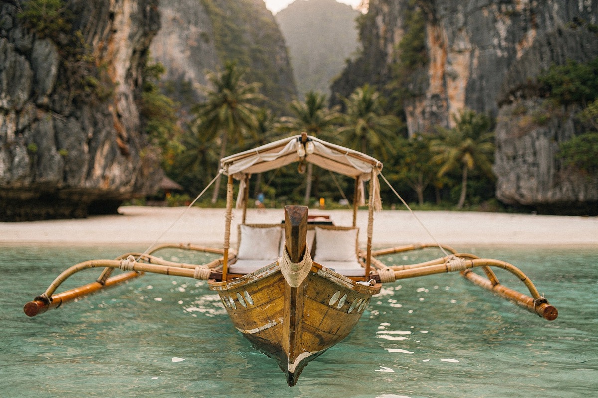 An empty wooden bangka seen head-on from the water, bamboo outrigger arms spread wide, a bamboo canopy frame over linen-cushioned benches, clear turquoise shallows, sheer karst walls either side.