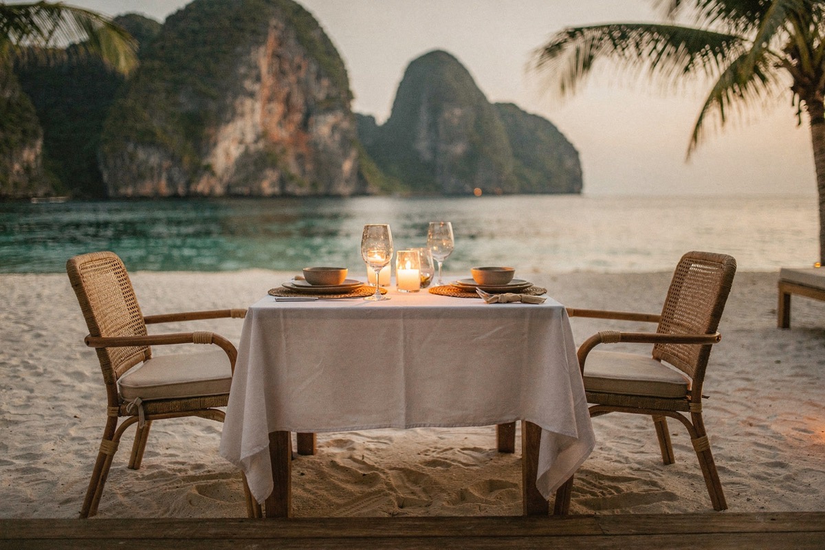 A table for two on white sand: white linen cloth, two place settings, wine glasses and a lit candle, two empty cane-backed armchairs, turquoise water and karst pinnacles, golden hour.