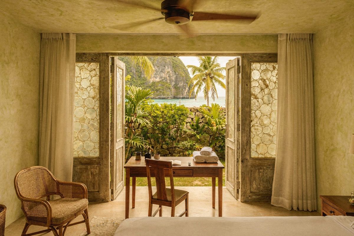 Lime-plaster walls in soft sage, capiz-shell panelled doors open onto a walled tropical garden, a teak writing desk and chair with folded towels, a ceiling fan above, karst and turquoise water beyond.