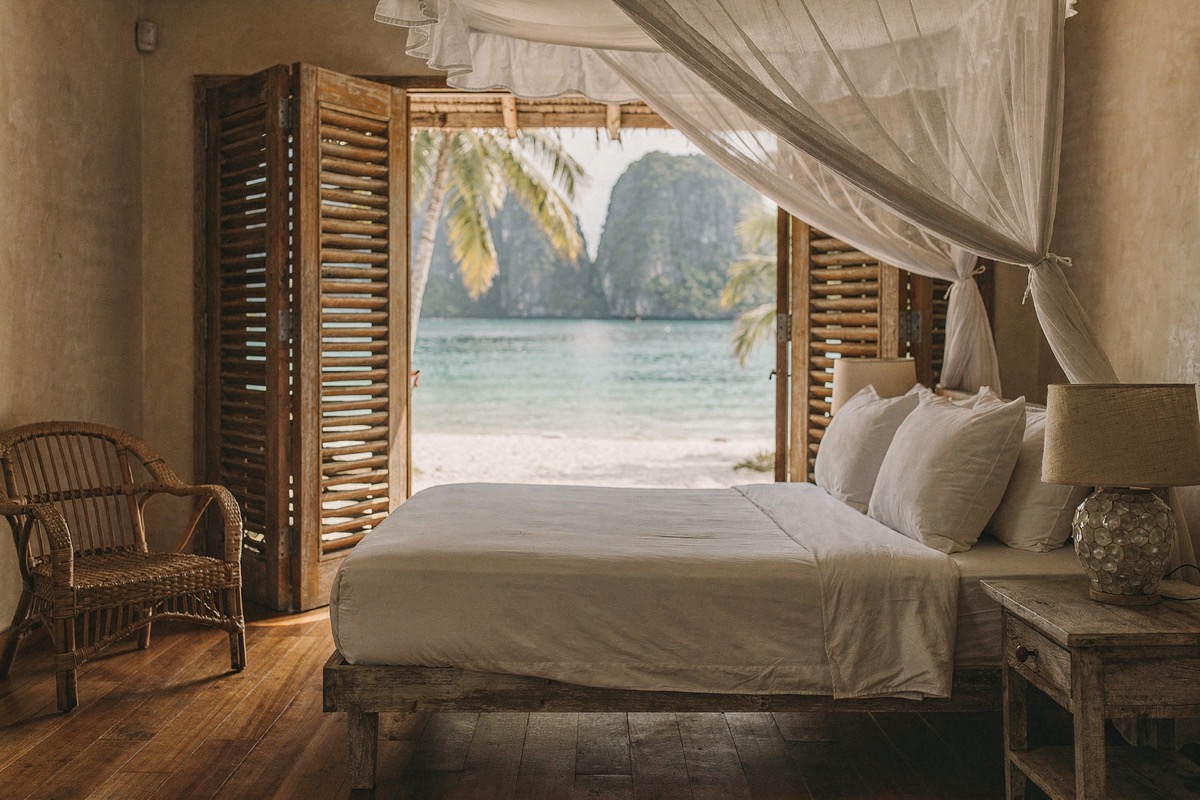 A low timber bed with turned-down white linen under a gathered mosquito net, timber louvred doors folded open to white sand and a turquoise lagoon, a rattan chair, wide-plank teak floor, morning light.