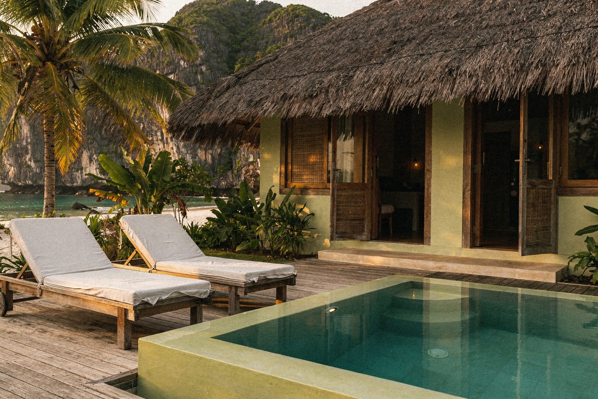 A private plunge pool with lime-plaster coping, two loungers on a timber deck, a thatched residence with open timber doors and a capiz-shell window, heliconia planting, karst and bay beyond, late afternoon.