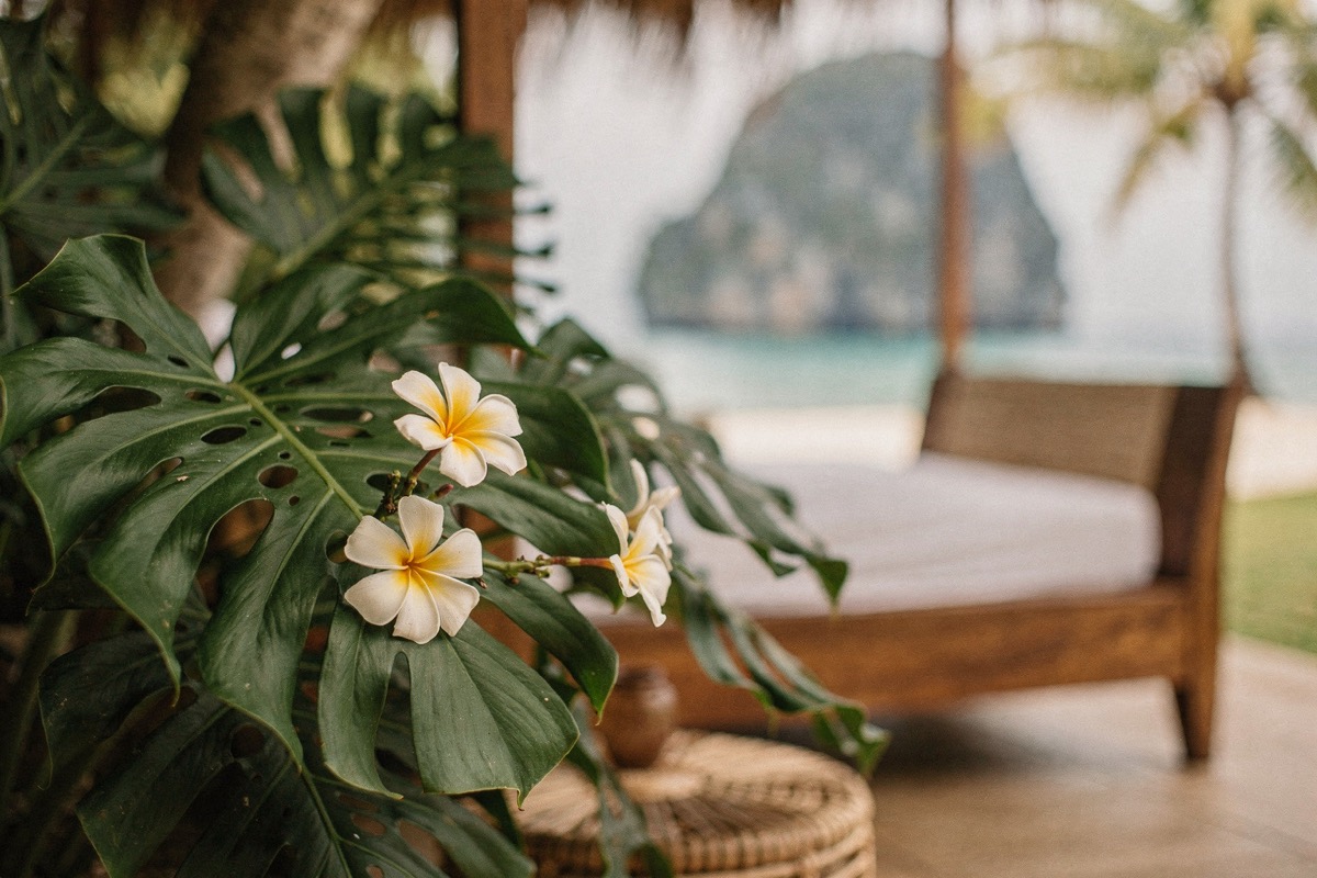 Monstera leaves and white frangipani flowers in sharp foreground focus, a blurred timber daybed, karst headland, palms and turquoise water behind, soft shade.
